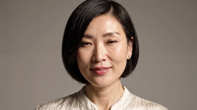 Confident middle aged Asian woman portrait on neutral studio background, calm female looking at camera, natural beauty lifestyle headshot