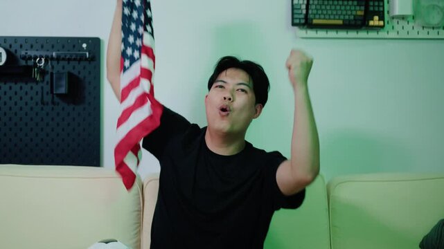 Stressed Asian Man Holding American Flag Watching Soccer Match on Sofa with Football Beside Him