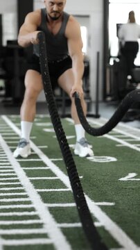Muscular athlete performing battle rope exercise in a modern gym. Functional strength training focused on endurance, power, cardio and full body fitness.