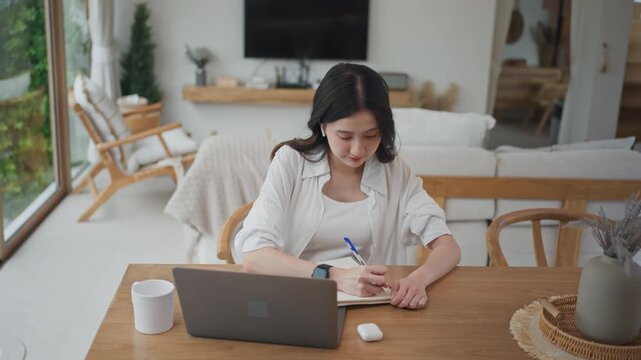 Asian woman writing notes in notebook while learning from online course on laptop at home, studying and planning tasks with focus in a bright modern living room