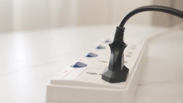 Person plugging black power cable into white extension socket strip indoors close up footage.