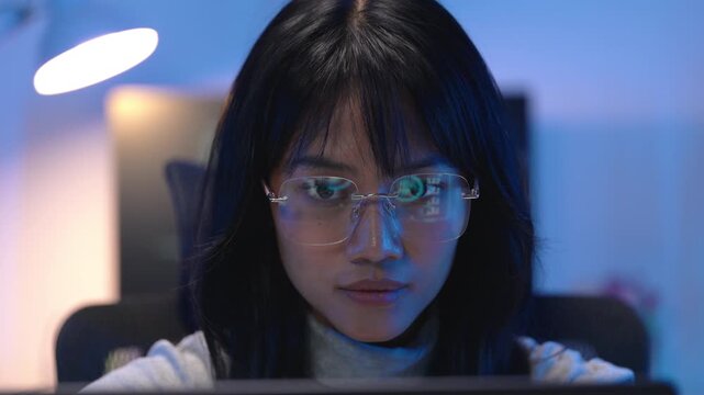 Asian woman wearing glasses working in cybersecurity, monitoring computer screen with blue light reflecting on her face, analyzing network security data late at night