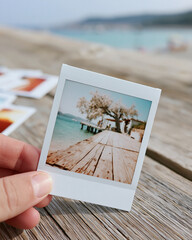 Fototapeta premium Hand holds instant photo of wooden pier. Tree stands near turquoise water. Soft focus blurs distant beach and sky. Coastal scene evokes calm, peaceful feeling. Ideal for vacation, travel