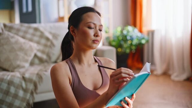 Thoughtful young asian woman writing fitness diary, planning workout notes home