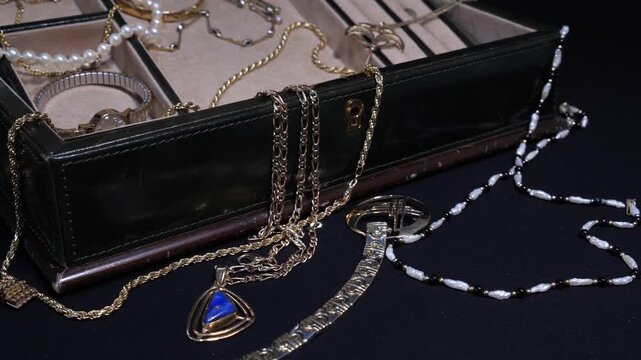 A close up cinematic shot of antique jewel box with shiny gold and pearl necklaces and other pieces of jewelry on black background.