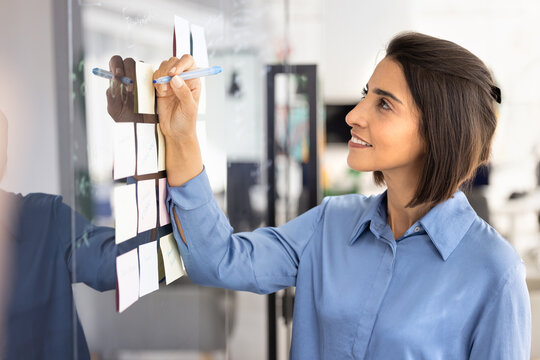 Businesswoman, office employee standing at glass board with attached paper sticky notes, holding marker, writing to-do list, planning ideas, organizing corporate tasks, creating visual business plan
