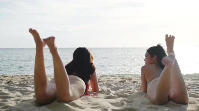 Attractive woman lies on beach near sea with female friend while sunbathing on warm sand during summer vacation, happy young women enjoy beautiful sunny day at tropical resort.