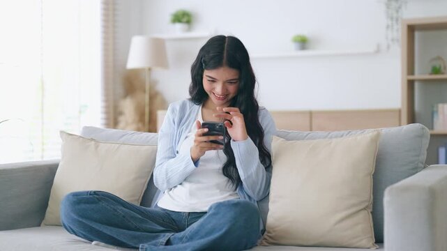 Happy young asian woman relax on comfortable couch at home texting messaging on smartphone, smiling girl use cellphone, chatting online message, shopping online from home