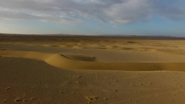 High angle aerial view captures wide desert plain with scattered small dunes and faint tracks across golden sand under clear daylight conditions.