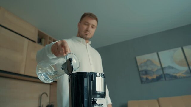 In the foreground, the focus is on a coffee maker into which a man in a white shirt pours water at home.