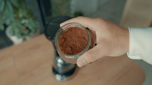 A hand with a can of ground coffee appears against the background of a blurred coffee maker and demonstrates it by shaking the can. The coffee symbol.