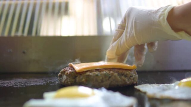 Chef hand placing slice of yellow cheddar cheese on juicy beef burger patty frying on hot commercial kitchen griddle.
