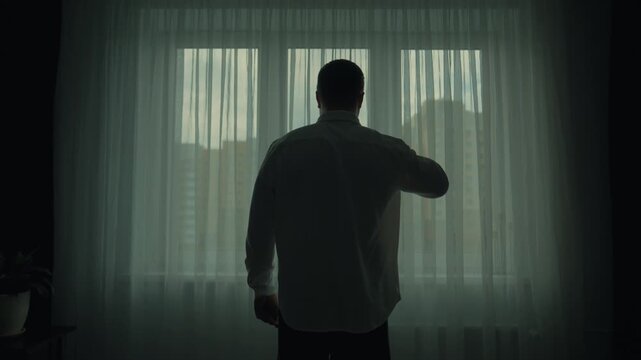 A man with a cup of coffee goes to the window, looks out and drinks coffee. Silhouette of a man drinking a drink by the window.