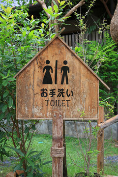 A rustic wooden sign with Japanese characters and clear symbols points the way to the restroom amidst a peaceful green garden.