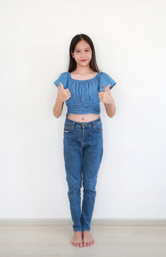 A smiling young Asian girl in a blue denim outfit standing against white wall, giving a double thumbs-up gesture to the camera.