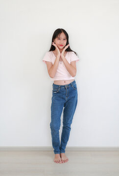 Charming young girl in pink wrap top and blue jeans posing playfully against white wall, framing her face with her hands.