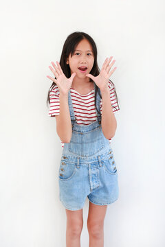 A cheerful young Asian girl wearing denim overalls and a striped t-shirt standing against a white background, raising both hands near her face with an excited, shouting expression.