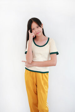Asian girl with long dark hair, in white and green trimmed t-shirt and yellow pants, stands confidently against white background, resting her chin on her hand in a thinking pose.