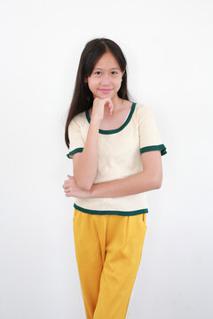 Cute Asian girl with long black hair, in a cream t-shirt with green trim and yellow pants, stands posing against white background, her hand on her chin, with pleasant smile.