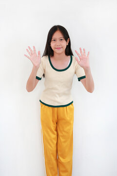 Smiling young Asian girl wearing cream t-shirt and yellow track pants standing against white wall while raising both of her hands to wave or show her palms.