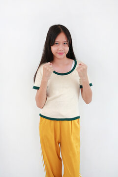 A young Asian girl in white t-shirt with green trim and yellow pants, standing against white background while raising her fists with a determined and cheerful expression.