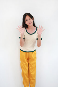 Happy young Asian girl in cream and green t-shirt and yellow pants standing against white background, smiling and waving both hands.