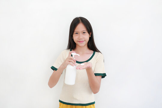 Smiling young Asian girl holding white pump bottle and applying moisturizing skin lotion onto her hand, isolated on white background.