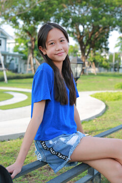 Smiling young Asian girl wearing a blue t-shirt and denim shorts sitting on a bench in a public park, looking towards the camera.