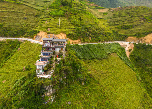 Aerial view of a building perched on a steep, green, terraced hillside overlooking the Nho Que River valley with a winding road in Meo Vac, Ha Giang, Vietnam.
