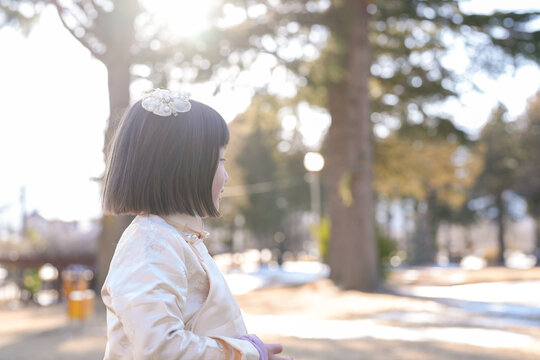 Young girl stands in sunlight, hair adorned with a small white flower. She gazes softly ahead, lost in quiet contemplation. Trees stand tall behind her, bathed in warm, golden light