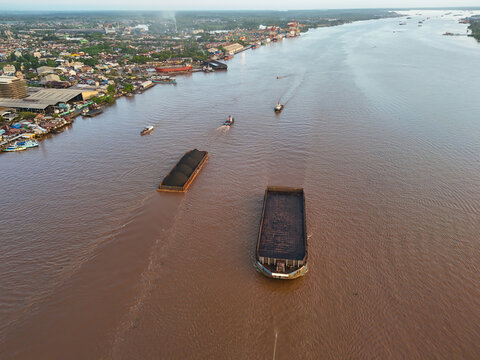 Aerial view of coal barges navigating the Barito River with industrial port facilities and city buildings along the banks in Banjarmasin, Kalimantan Selatan, Indonesia.