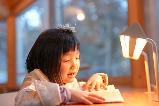 Soft light warms a quiet reading moment. Child leans forward, eyes focused on pages. Warm lamp casts gentle glow beside her. Cozy wooden table holds open book