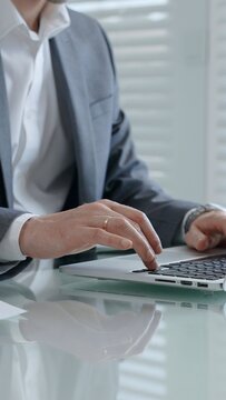 Professional hands typing on a laptop keyboard over a reflective glass desk. Showing a focused person working remotely from an office or home setup with blinds in the background