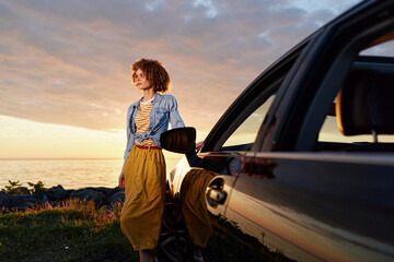 Naklejka premium Smiling woman by a car at sunset, standing on a coastal cliff in a denim shirt and yellow skirt, radiating relaxed lifestyle and confident calm.