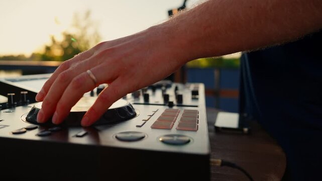 dj playing music adjusting controls on outdoor setting with trees and sunset in the background, focused and energetic close-up shot of hands on wooden surface of dj mixer.