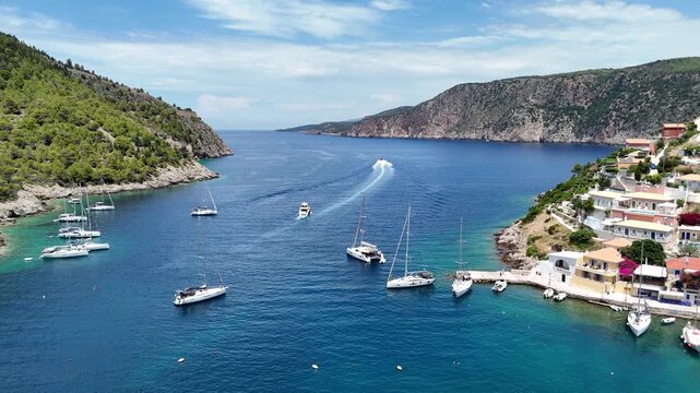 Aerial drone video of Assos village in Kefalonia, Greece, with colorful waterfront buildings, anchored boats, turquoise waters, and scenic Mediterranean coastal views