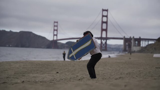 Man Launching Painted Board Overhead on Beach Near Golden Gate Bridge Against Strong Wind