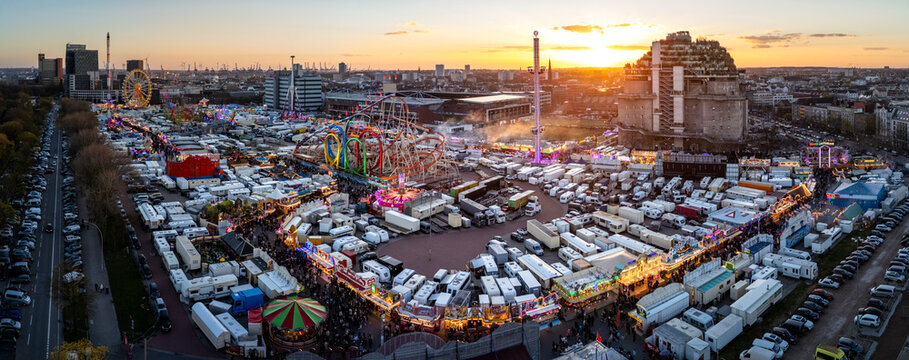 Hamburg, Germany - 01 April 2026: Aerial view of Heiligengeistfeld during the Hamburger Dom fair, featuring roller coasters, carnival rides, and numerous trailers under a golden sunset sky.