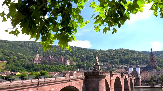 Green tree leaves frame Alte Br&uuml;cke Karl Theodor Bridge with Heidelberg Castle, old town buildings and hills on a bright summer day.