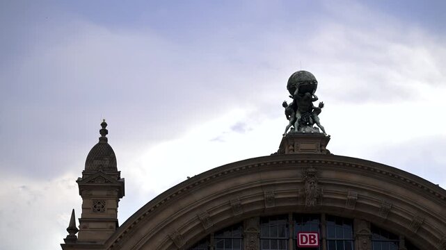 Atlas sculpture crowns the arched entrance of Frankfurt Main Hauptbahnhof beneath a cloudy sky with the DB logo visible.