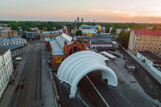 Riga, Latvia - 06 June 2019: Aerial view of Agenskalns Market featuring its red brick architecture and a large white inflatable arch structure, surrounded by urban streets during the golden hour.