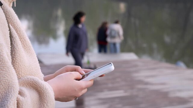Person holds a phone, focused on screen. Background shows blurred pedestrians near water. Calm, everyday moment of digital interaction. Ideal for scenes of modern connectivity or urban leisure