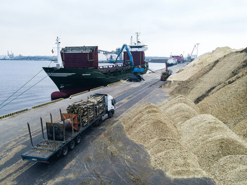 Riga, Latvia - 03 November 2021: Aerial view of a cargo ship being loaded with wood chips at the port, with a truck carrying timber and large piles of wood chips on the dock.
