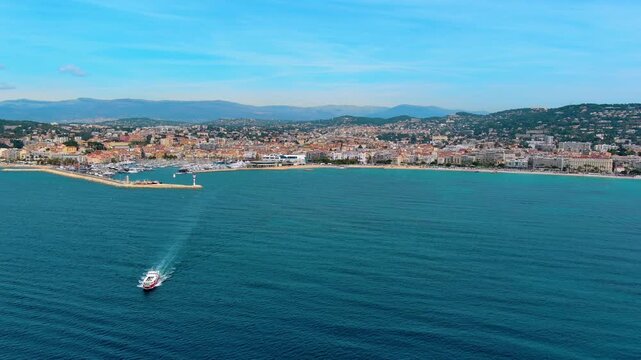 Aerial view of a passenger ferry sailing off Cannes, France, toward the old port with yachts, the Palais des Festivals, La Croisette beach, Le Suquet old town and Alpine foothills behind.