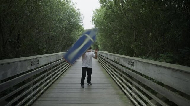 Man Quickly Spinning Blue Sign on Wooden Footbridge Surrounded by Trees - Steady Cam Forward