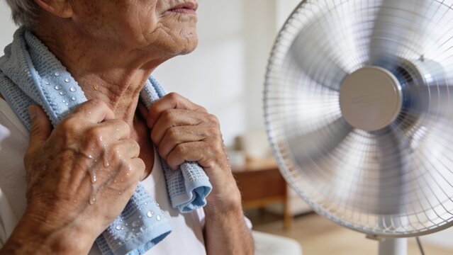 Senior person cooling down with a towel in front of an electric fan during a summer heatwave at home.