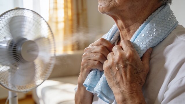 Senior person cooling down with an ice pack around the neck beside an electric fan during a hot summer day.