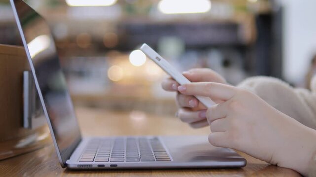 Hands use smartphone beside laptop in cozy cafe setting. Person focuses intently on phone screen. Soft background lighting creates warm, relaxed mood. Ideal for remote work, digital communication