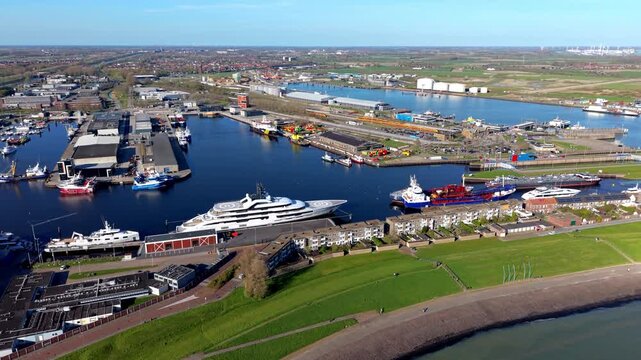 Aerial wide shot of a bustling seaport harbor channel filled with large luxury yachts, commercial ships, and cargo barges next to a coastal town on a bright sunny day