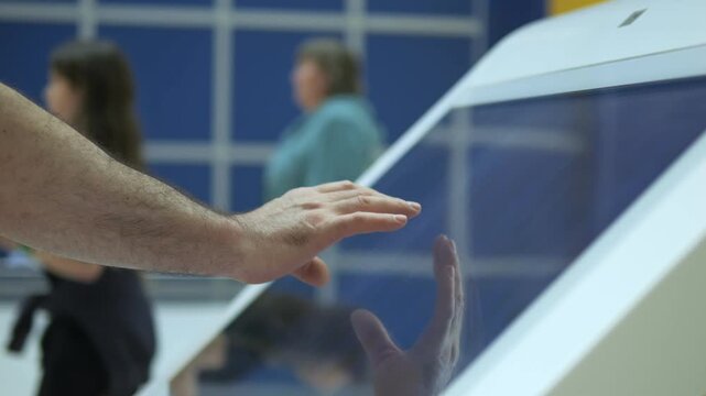 Hand interacts with modern touch screen device. Fingers glide across glass panel. Background shows blurred indoor setting with people moving. Scene conveys technology, interaction